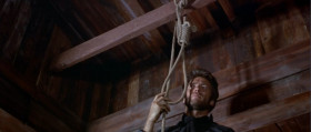 Now make sure the rope is tight. It's got to hold the weight of a pig. Now put the rope around your neck. That's very good. It's too big for your neck, huh? We fix that right away. I have another system. A little different than yours. I don't shoot the rope, I shoot the legs off the stool.