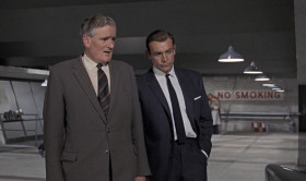 - Morning, Q.
- Good morning, 007. This way, please. My, we are busy this morning. It's not perfected yet.
- Where's my Bentley?
- Oh, it's had its day, I'm afraid.
- But it's never let me down.
- M's orders, 007. You'll be using this Aston Martin D85, with modifications. Now, pay attention, please. Windscreen, bulletproof. As are the side and the rear windows. Revolving number plates, naturally. Valid all countries. Here's a nice little transmitting device called a homer. You prime it by pressing that back like this. You see? The smaller model is now standard field issue, to be fitted into the heel of your shoe. Its larger brother is magnetic. Right. To be concealed in the car you're trailing while you keep out of sight. Reception on the dashboard here. Auto-visual. Range, 150 miles.
- Ingenious. And useful, too. Allow a man to stop off for a quick one en route.
- It has not been perfected after years of patient research entirely for that purpose, 007. And incidentally, we'd appreciate its return, along with all
your other equipment, intact, for once, when you return from the field.