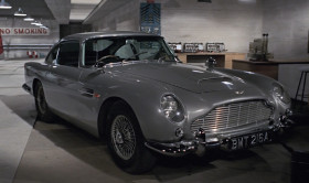 - Morning, Q.
- Good morning, 007. This way, please. My, we are busy this morning. It's not perfected yet.
- Where's my Bentley?
- Oh, it's had its day, I'm afraid.
- But it's never let me down.
- M's orders, 007. You'll be using this Aston Martin D85, with modifications. Now, pay attention, please. Windscreen, bulletproof. As are the side and the rear windows. Revolving number plates, naturally. Valid all countries. Here's a nice little transmitting device called a homer. You prime it by pressing that back like this. You see? The smaller model is now standard field issue, to be fitted into the heel of your shoe. Its larger brother is magnetic. Right. To be concealed in the car you're trailing while you keep out of sight. Reception on the dashboard here. Auto-visual. Range, 150 miles.
- Ingenious. And useful, too. Allow a man to stop off for a quick one en route.
- It has not been perfected after years of patient research entirely for that purpose, 007. And incidentally, we'd appreciate its return, along with all
your other equipment, intact, for once, when you return from the field.